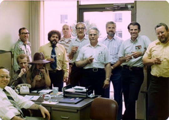 This was taken in '76 or '77 at the small industrial office that was rented for the development of the Reflex disk drive product. It was about a mile or two south on Redhill from the Microdata main plant and generally referrd to as the "skunk works". Most of these guys had been hired away from Century Data (Calcomp) for this project. The photo was taken at a party celebrating some milestone... I think perhaps the first working prototype. Seated at the lower left: Vice President Bud Bleininger Seated behind the desk: Bill Helpard, with Helen Samarin on his lap. Standing, from left to right: Jerry Dischino, Mike Clark, Jim Duke, Bob Gertz, Frank Grosch, Bill Zeissner, Steve Klinger, and Chet Kulma. (Photo courtesy John Van Valkenburgh - Microdata '76-'81).
